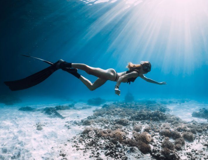 Woman freediver with fins swim over sandy sea and sun rays. Underwater ocean